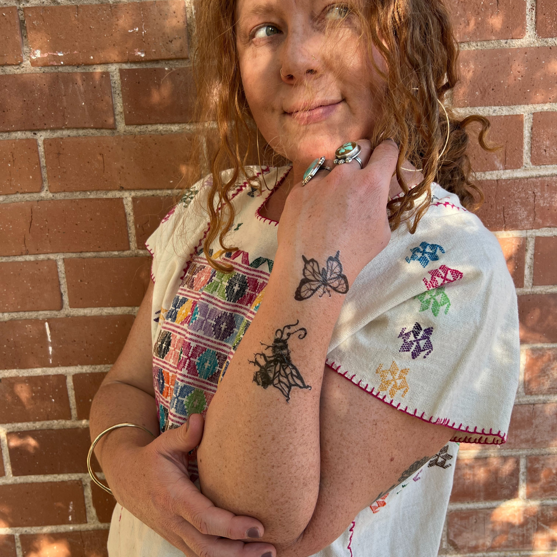 woman standing against brick wall, with temporary tattoos of a butterfly and bumble bee on forearm. 