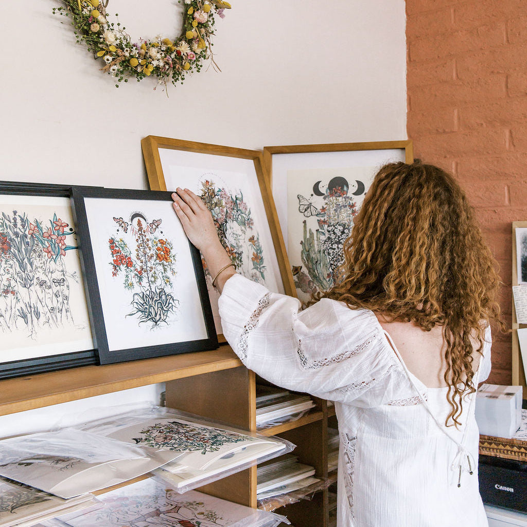Woman arranging framed artwork on a shelf with a floral wreath on the wall.