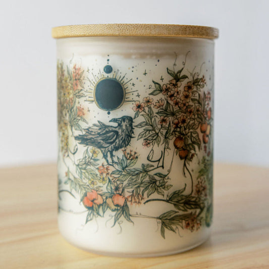 A glass jar candle with a bamboo lid, featuring a vintage-style illustration of a raven and botanical elements on a cream-colored wax, set on a light wooden surface against a white background.