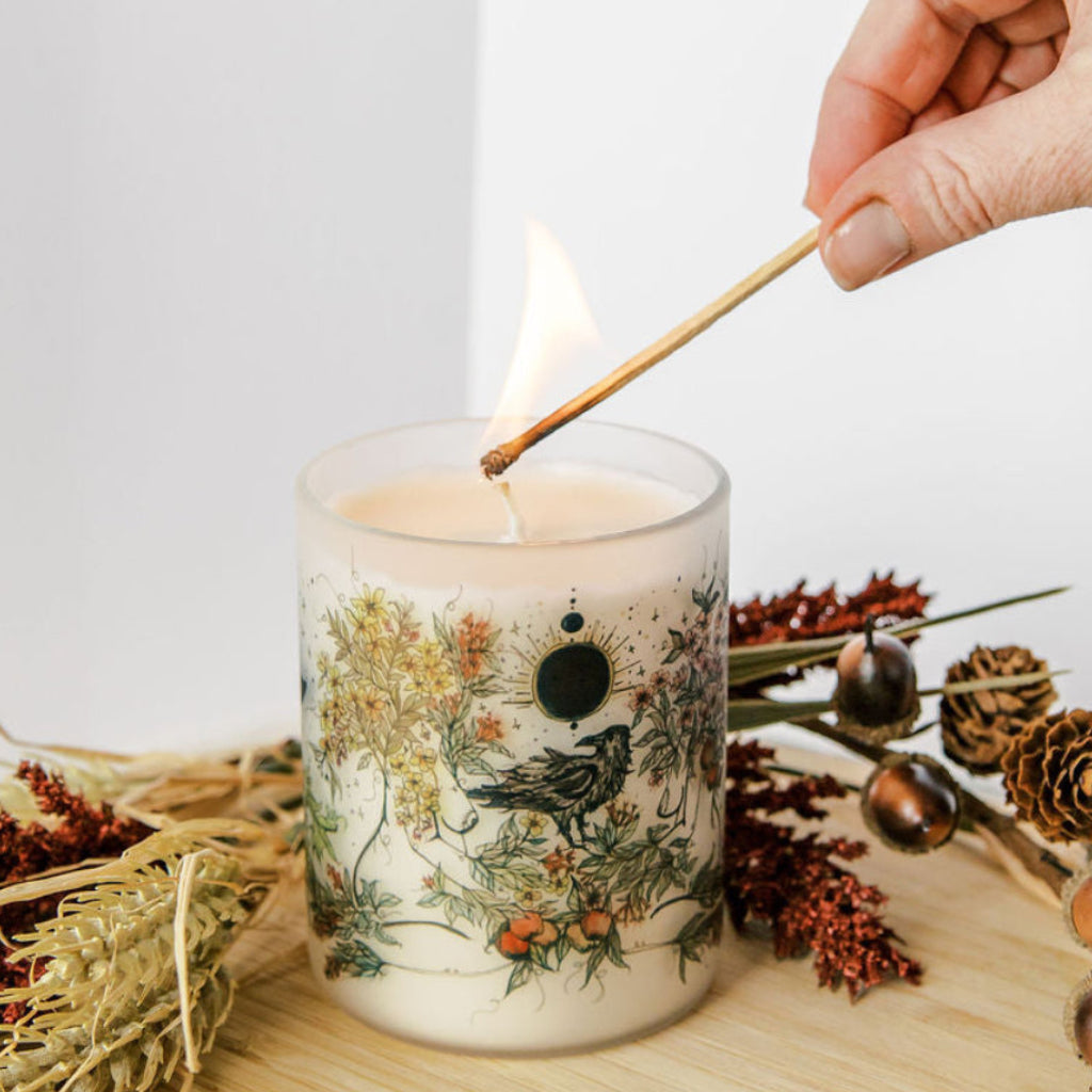 hand lighting mabon fall equinox soy candle with botanical artwork wrapped around frosted glass jar by marcy ellis in tucson az
