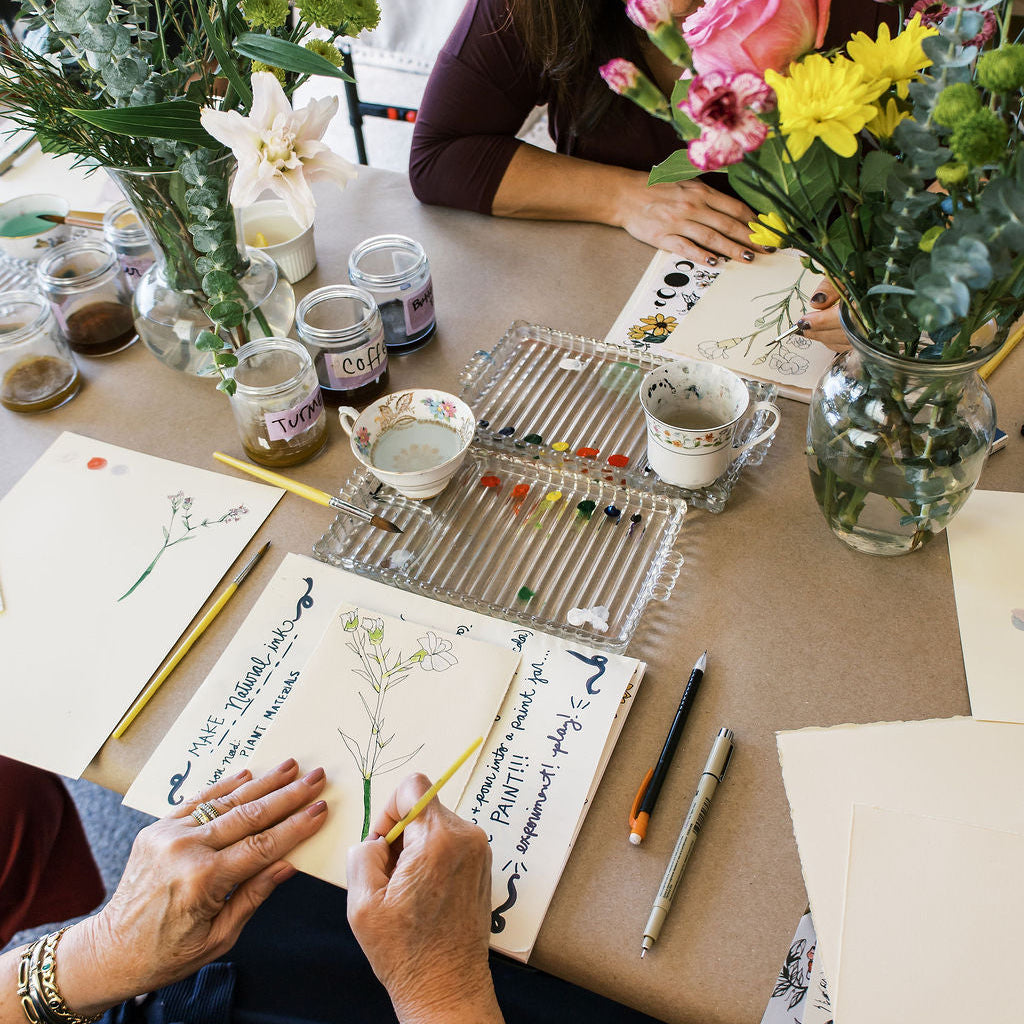 Two people working on art projects at a table with supplies and flowers.