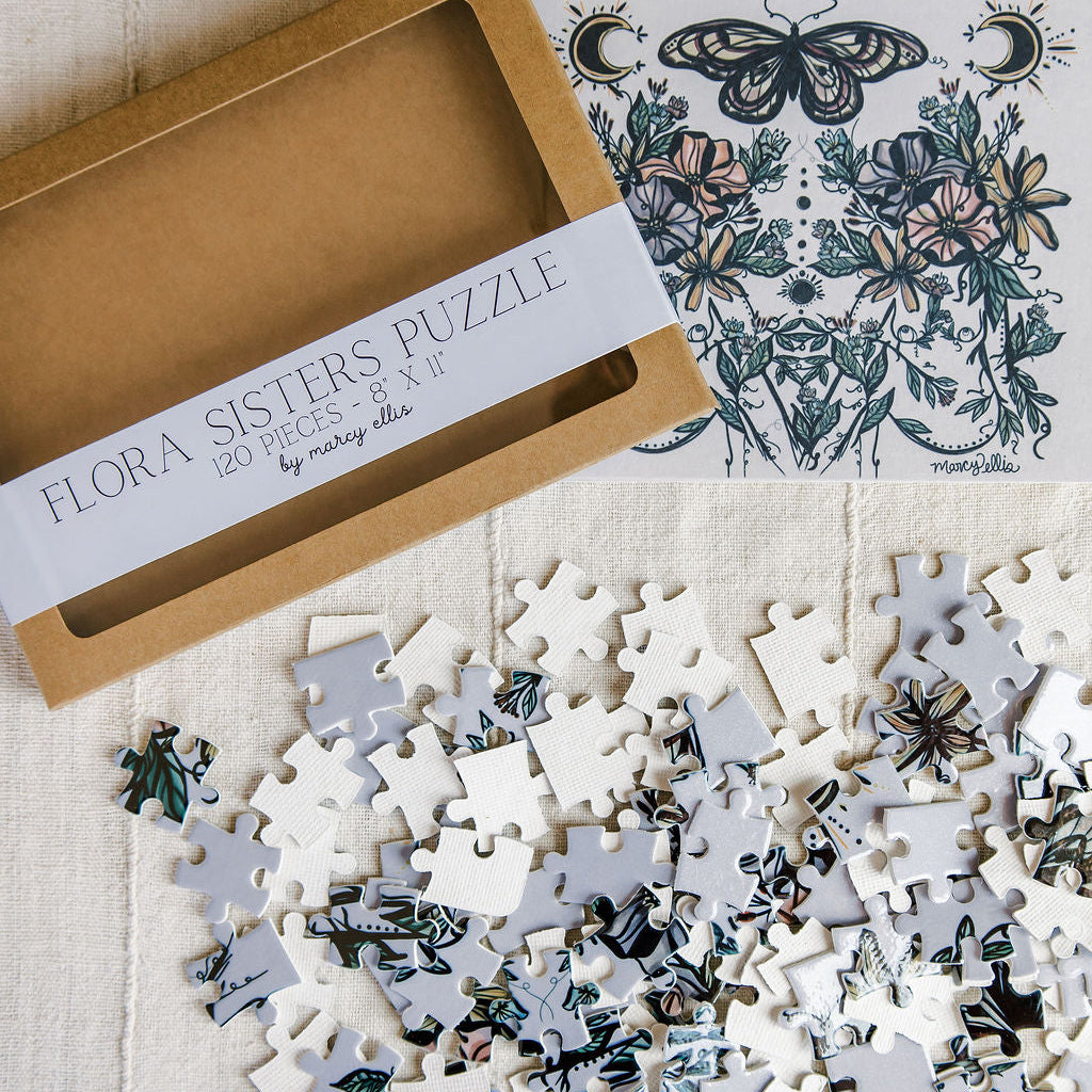 Flora Sisters Puzzle by Marcy Ellis, featuring a kraft paper box, the illustrated puzzle cover, and a scattered pile of white puzzle pieces with botanical and floral designs on a textured cream fabric background.