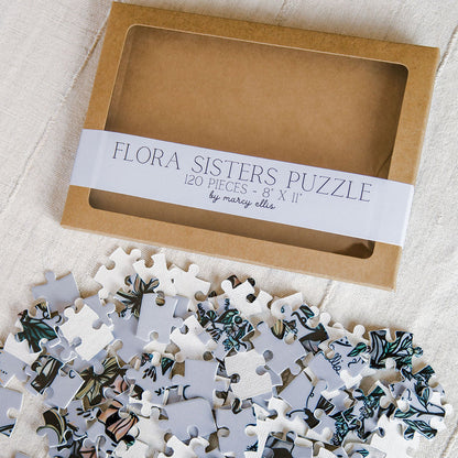 Flora Sisters 8x10 inch jigsaw puzzle by Marcy Ellis, shown with its brown cardboard packaging box and a pile of 120 patterned puzzle pieces on a textured white fabric background.