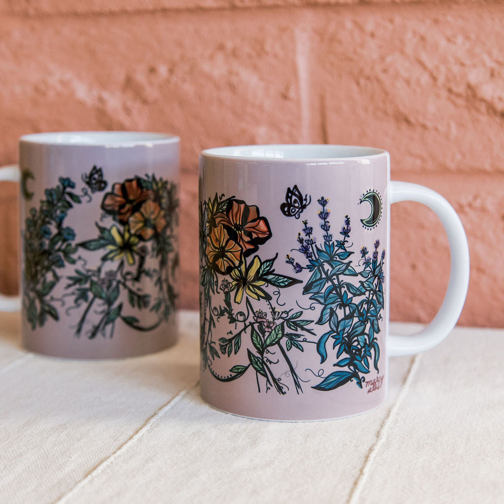 Two ceramic mugs by Marcy Ellis with a lavender background and botanical figure illustrations, displayed on a light wooden surface against a terracotta brick wall.