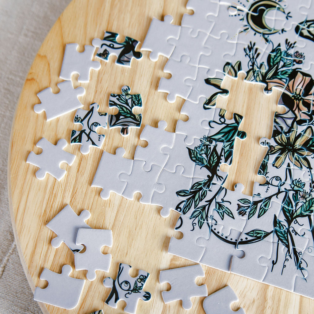 A partially completed botanical feminine  and celestial jigsaw puzzle by Marcy Ellis with white pieces, displayed on a round light wood board.