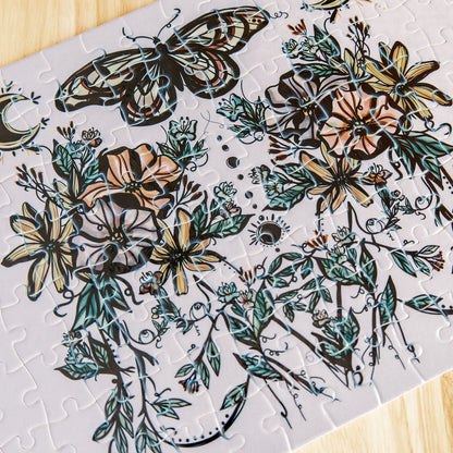 A completed jigsaw puzzle featuring a botanical floral feminine design with a butterfly, flowers, leaves, and crescent moons by Marcy Ellis placed on a light wood surface.