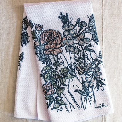 A folded white waffle-weave tea towel with a botanical print of women intertwined with roses, flowers, and butterflies.
