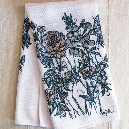 A folded white waffle-weave tea towel with a botanical print of women intertwined with roses, flowers, and butterflies.