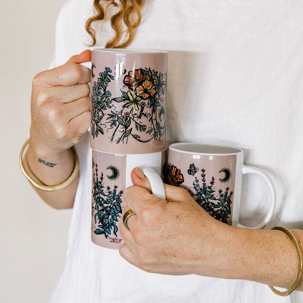 Marcy Ellis holds three ceramic mugs decorated with botanical illustrations, butterflies, and celestial motifs like crescent moons and stars. The mugs have a dusty pink background with dark green foliage and orange butterfly accents.