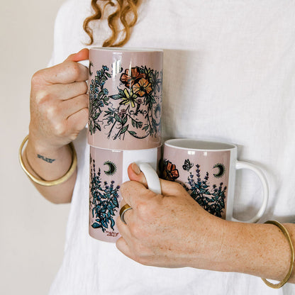 Marcy Ellis holds three ceramic mugs decorated with botanical illustrations, butterflies, and celestial motifs like crescent moons and stars. The mugs have a dusty pink background with dark green foliage and orange butterfly accents.