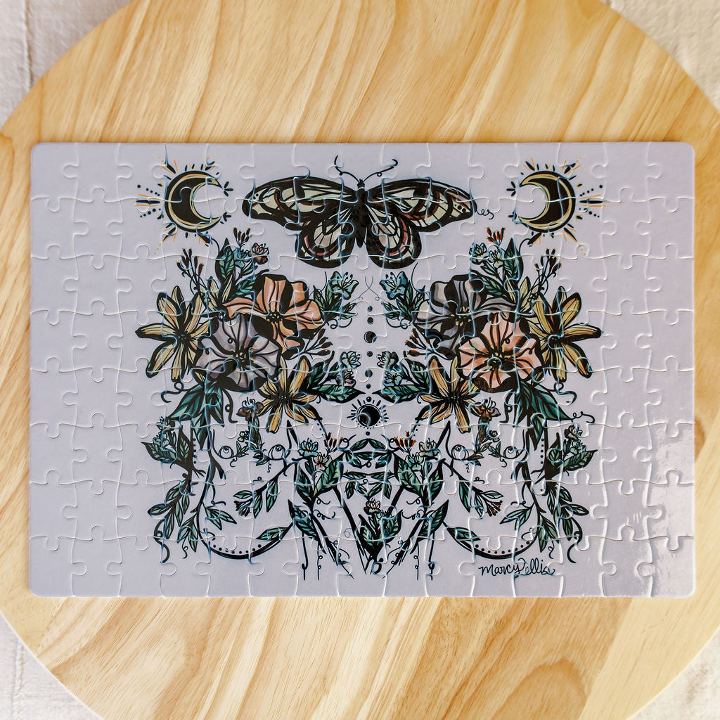 A completed jigsaw puzzle by Marcy Ellis featuring two symmetrical women with  florals growing and butterfly design on a pale background, placed on a round light wood surface.