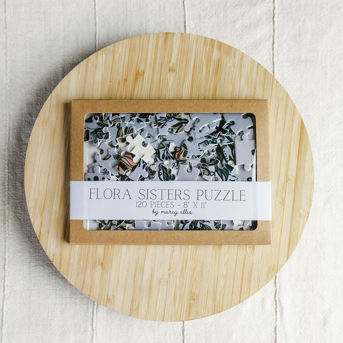 Flora Sisters Puzzle by Marcy Ellis, a 120-piece jigsaw puzzle in a light wood box with a clear window, displayed on a round bamboo board over a white wood background.