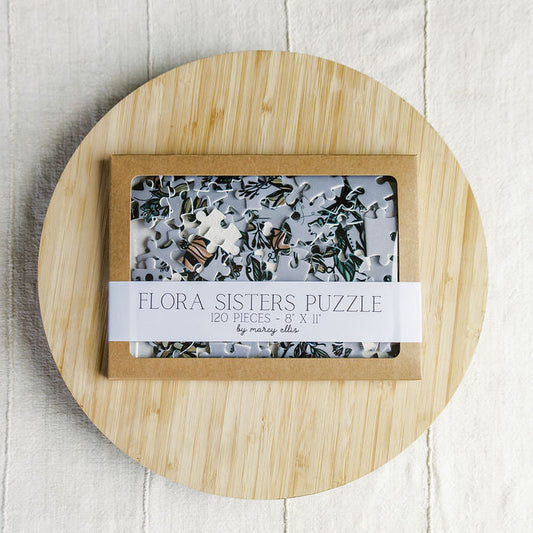 Flora Sisters Puzzle by Marcy Ellis, a 120-piece jigsaw puzzle in a light wood box with a clear window, displayed on a round bamboo board over a white wood background.