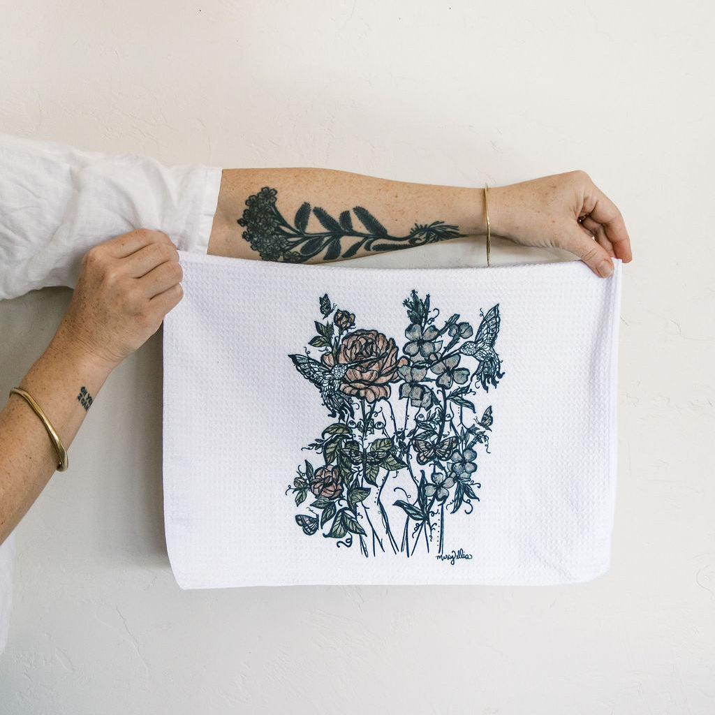 Marcy Ellis holds up a white kitchen towel with a detailed botanical floral print in dark teal and muted pink, against a plain off-white wall.