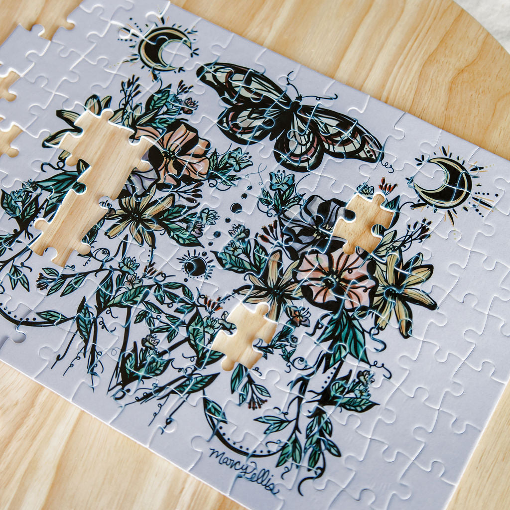 A partially assembled jigsaw puzzle by Marcy Ellis featuring two botanical and celestial women with a butterfly, flowers, leaves, and crescent moons on a pale background, placed on a light wood surface.