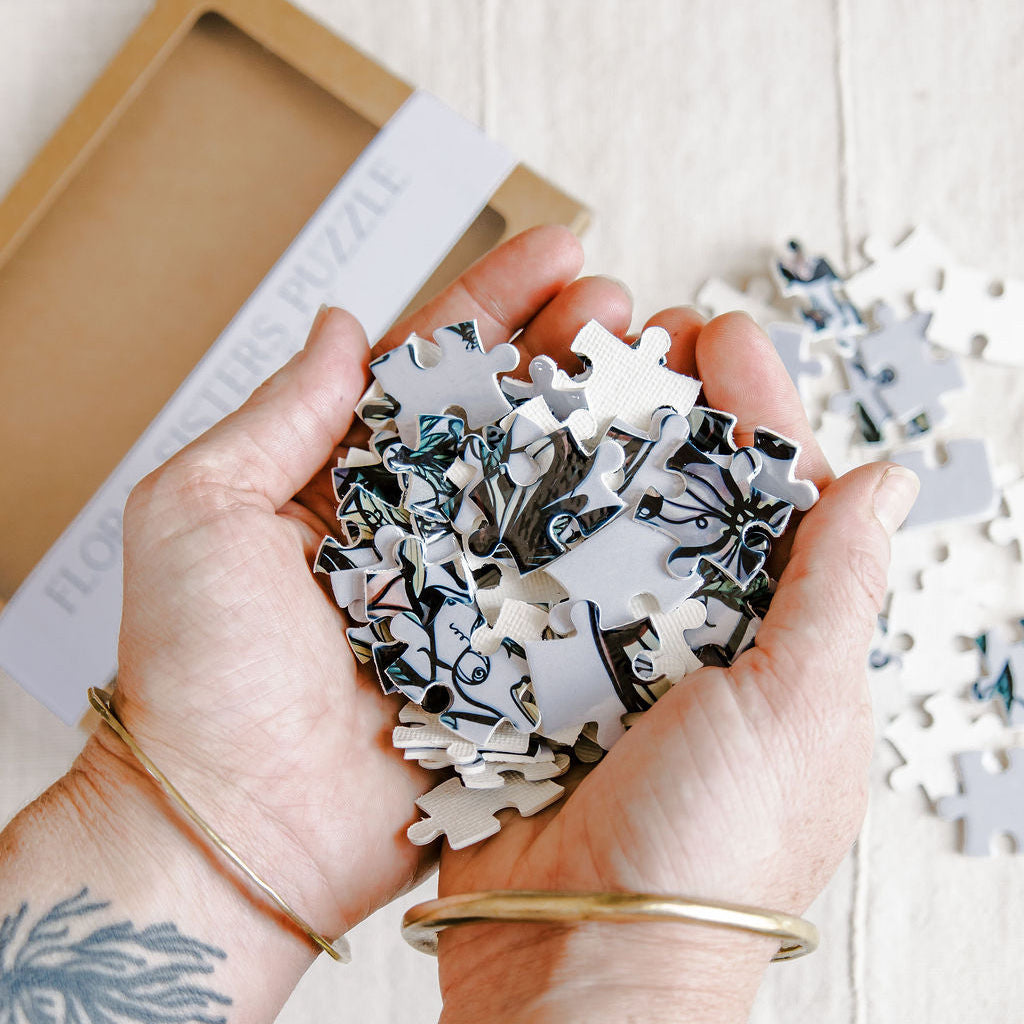 Marcy Ellis holding a pile of white jigsaw puzzle pieces with black and blue botanical line art, with a partially opened kraft paper box in the background on a light wood surface.