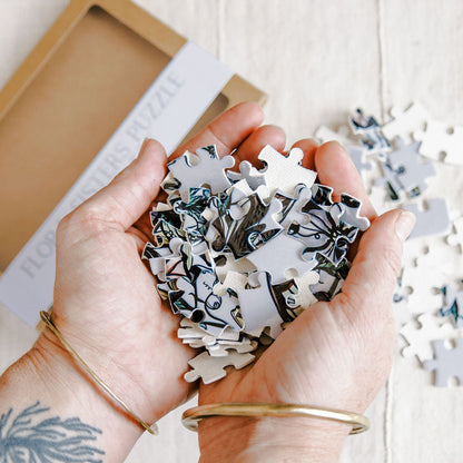 Marcy Ellis holding a pile of white jigsaw puzzle pieces with black and blue botanical line art, with a partially opened kraft paper box in the background on a light wood surface.