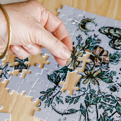 A close-up of a hand placing a flora sisters Marcy Ellis puzzle piece onto a partially assembled botanical jigsaw puzzle on a light wood surface.
