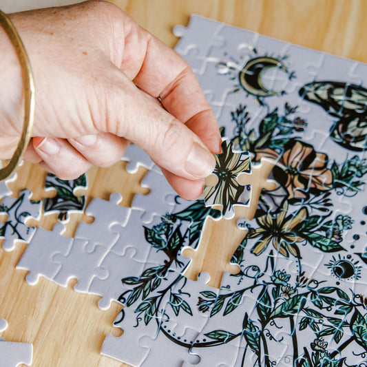 A close-up of a hand placing a flora sisters Marcy Ellis puzzle piece onto a partially assembled botanical jigsaw puzzle on a light wood surface.