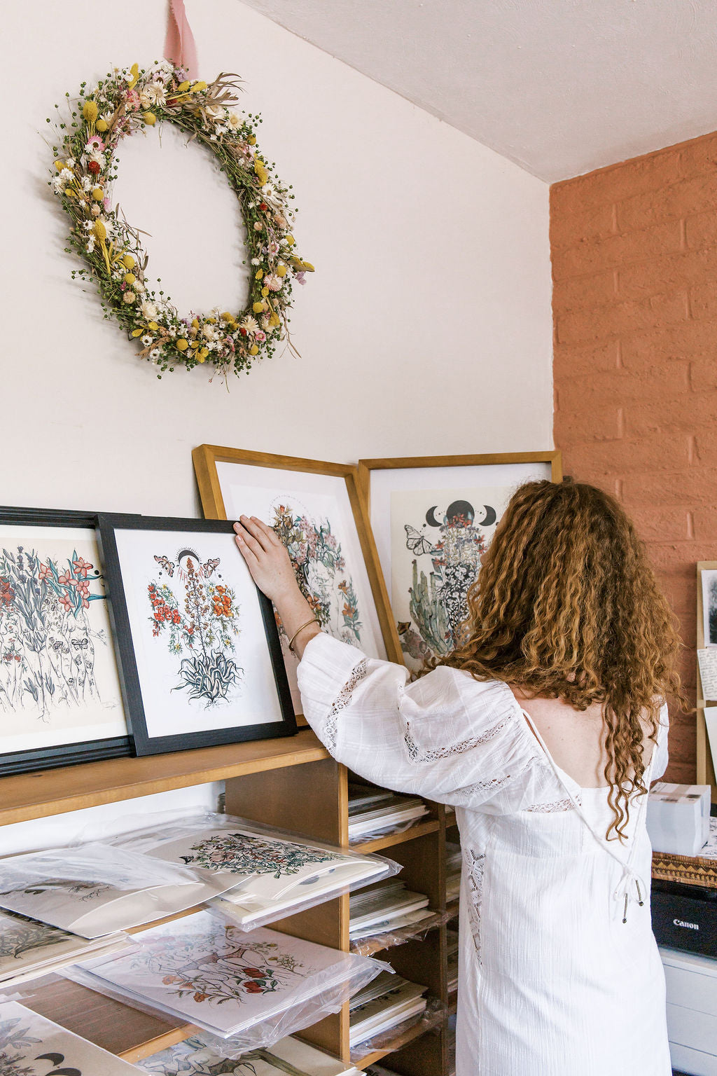 Woman arranging framed artwork on a shelf with a floral wreath on the wall.