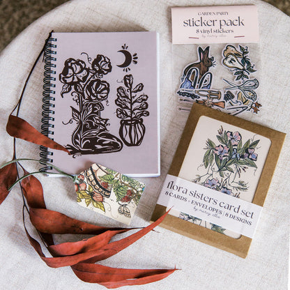 Stationery items including a notebook, cards, and stickers on a textured surface with leaves.