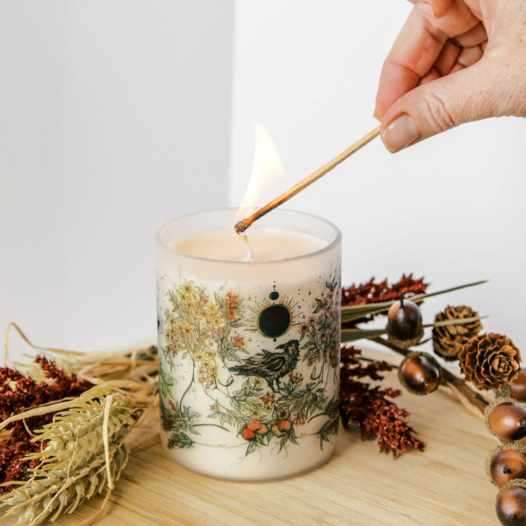 hand lighting mabon fall equinox soy candle with botanical artwork wrapped around frosted glass jar by marcy ellis in tucson az
