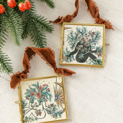 Two square gold-framed ornaments with rustic brown ribbon hangers, featuring Yule feminine botanical illustrations by marcy ellis, displayed on a textured cream fabric next to pine branches and red berries.