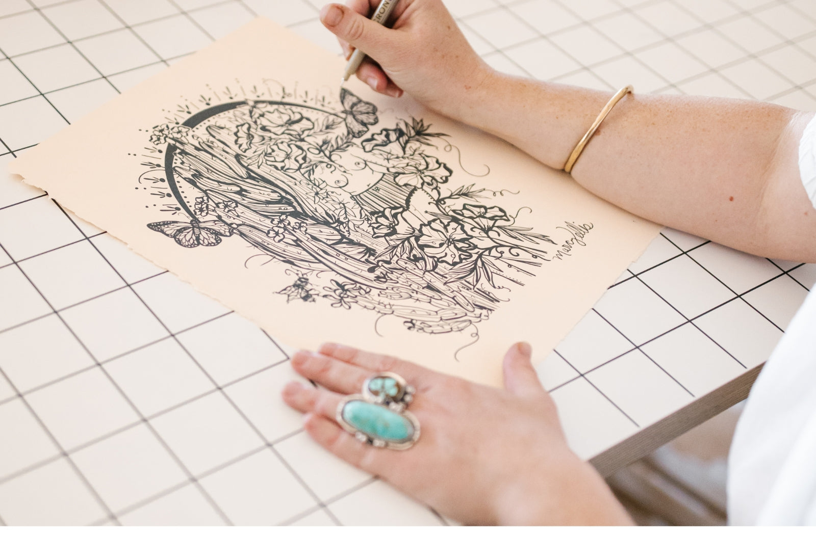 Person using a craft knife to draw a figure in a desert scene with black and  white illustration on beige paper.