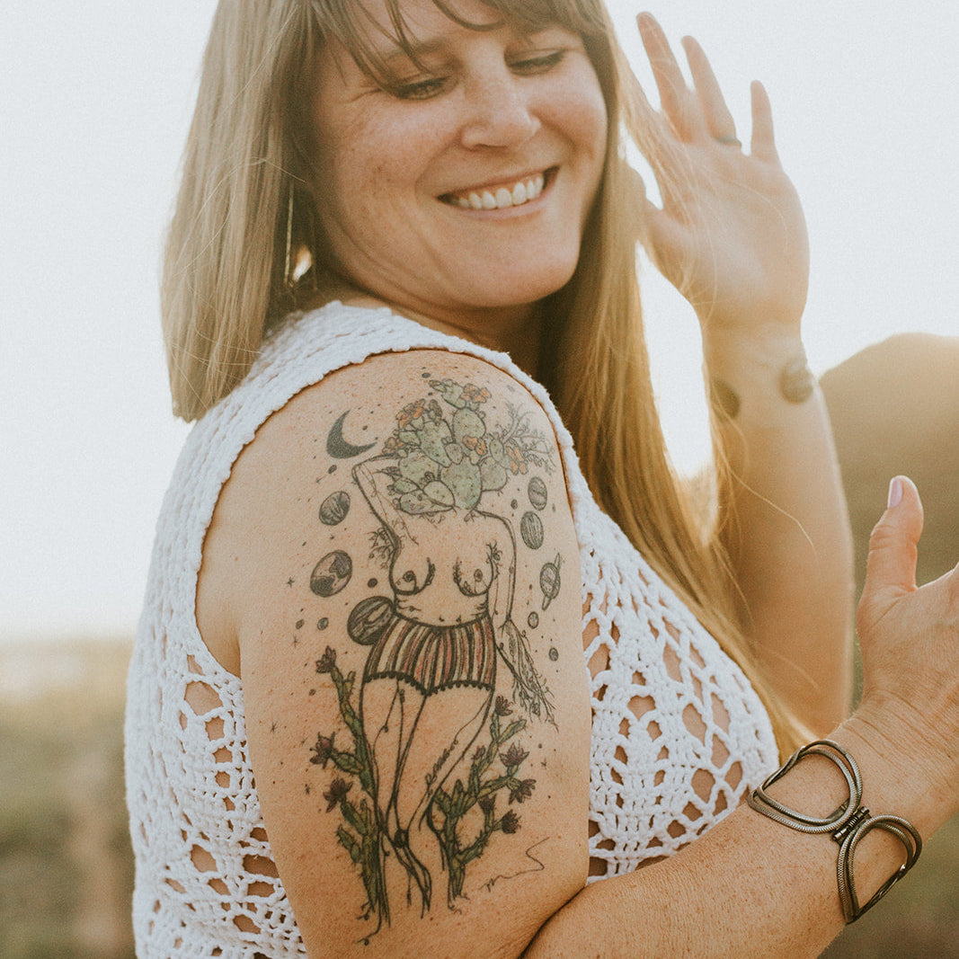 A smiling woman with a large colorful tattoo on her upper arm of marcy ellis's botanical women body, wearing a white crochet top, photographed outdoors in warm natural light.