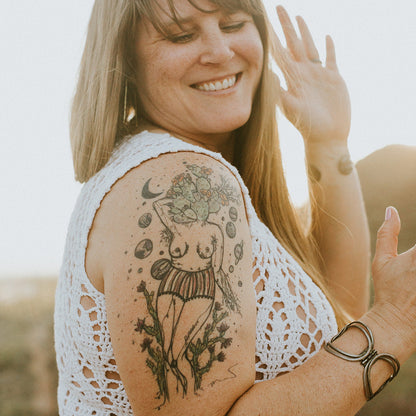 A smiling woman with a large colorful tattoo on her upper arm of marcy ellis's botanical women body, wearing a white crochet top, photographed outdoors in warm natural light.
