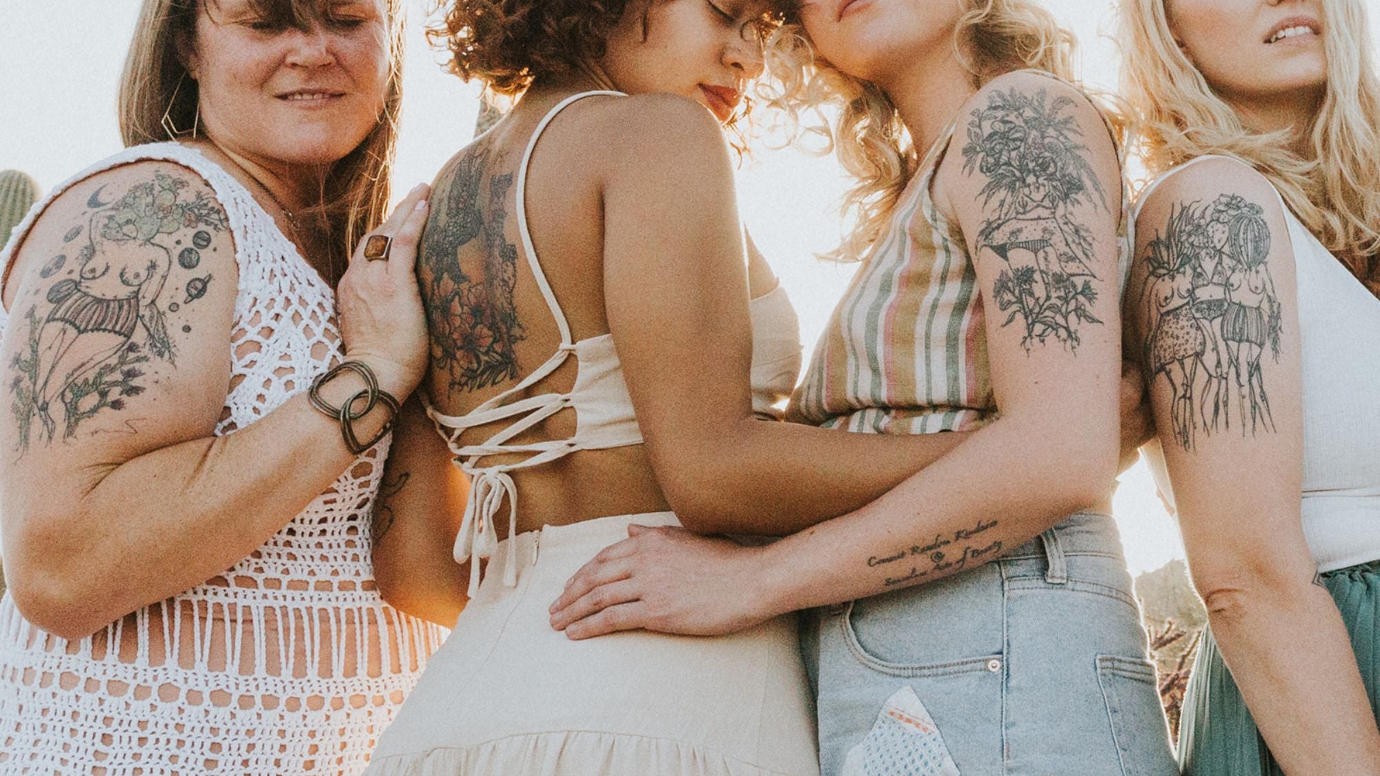 Four women embracing each other with Marcy Ellis tattoo designs , showcasing various black ink tattoos on their arms, backs, and chests. They are wearing casual bohemian-style tops and jeans under warm, bright sunlight.
